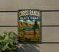 A metal sign for Cross Ranch State Park in North Dakota, featuring a scenic landscape with mountains, a lake, and a pine tree.