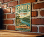 A vintage metal sign for Crowder State Park in Missouri, featuring a scenic landscape with a lake and mountains, is displayed on a brick wall.