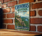 A vintage metal sign for Crowders Mountain State Park in North Carolina, featuring a scenic mountain landscape with a river and pine trees.