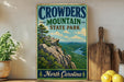 A vintage-style sign for Crowders Mountain State Park in North Carolina, featuring a scenic mountain landscape with a river and pine trees.