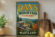 A vintage metal sign for Dan's Mountain State Park in Maryland, featuring a scenic mountain landscape with pine trees and rocky outcroppings.