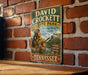 A vintage metal sign for David Crockett State Park in Tennessee, featuring an illustration of a man with a rifle and a scenic landscape.