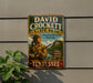 A vintage metal sign for David Crockett State Park in Tennessee, featuring a man in a fur-trimmed hat holding a rifle and standing in front of a scenic landscape.