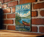 A vintage-style metal sign for Day Pond State Park in Connecticut, featuring a scenic image of a lake with a canoe, surrounded by trees and mountains.