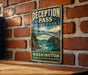 A vintage-style metal sign for Deception Pass State Park in Washington, featuring a scenic illustration of a bridge over a river surrounded by mountains.