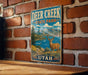 A vintage metal sign for Deer Creek State Park in Utah, featuring a deer standing in a scenic landscape with mountains, water, and trees.