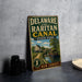 A vintage metal sign for Delaware and Raitan Canal State Park, featuring an illustration of a canal and a house, is displayed on a gray countertop next to a stack of black camera lenses.