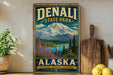 A vintage-style sign for Denali State Park in Alaska, featuring a mountain landscape with pine trees and a lake.
