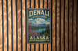 A vintage metal sign for Denali State Park in Alaska, featuring a mountain landscape with pine trees and a lake.