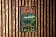 A vintage-style metal sign for Devil's Millhopper Geological State Park in Florida, featuring a scenic illustration of a bridge and palm trees.
