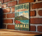 A vintage-style metal sign with the text "DIAMOND HEAD STATE MONUMENT HAWAII" and a tropical landscape depicting a mountain, palm trees, and a beach.
