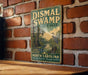 A vintage-style metal sign for DisMAL SWAMP State Park in North Carolina, featuring a scenic illustration of a swamp landscape with a bird in the foreground.