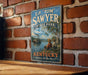 A vintage-style metal sign for E.P. Sawyer State Park in Kentucky, featuring a river scene with a steamboat and two people sitting on the riverbank.