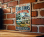 A vintage metal sign with the text "Welcome to East Tawas City Park" and an illustration of a gazebo and a lake, mounted on a brick wall.