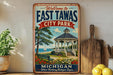 a vintage-style sign for East Tawas City Park, with a gazebo and a scenic view of the lake and mountains in the background.
