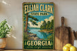 A vintage-style sign for Elijah Clark State Park in Georgia, featuring a scenic landscape with a river and pine trees.