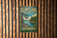 A vintage metal sign for Eljah Clark State Park in Georgia, featuring a scenic landscape with a river and pine trees.