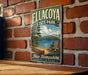A vintage metal sign for Ellacoya State Park, featuring a scenic image of a lake, mountains, and a red boat, is displayed on a brick wall.
