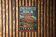 A vintage-style metal sign for the Enchanted Rock State Natural Area in Texas, featuring a desert landscape with a large rock formation and desert plants.