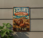 A vintage metal sign for Escalante Perfumed Forest State Park in Utah, featuring a scenic image of a forest with large logs and mountains in the background.