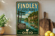 A vintage-style sign for Findley State Park in Ohio, featuring a scenic landscape with a river and trees, is displayed on a shelf next to a wooden cutting board and some lemons.