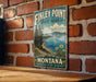 A vintage-style metal sign for Finley Point State Park in Montana, featuring a scenic landscape with mountains, pine trees, and a lake with flowers.