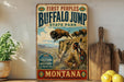 A vintage-style metal sign for Buffalo Jump State Park in Montana, featuring an illustration of a buffalo jumping over a cliff with Native American figures in the background.