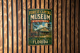 A vintage-style metal sign for the Forest Capital Museum in Florida, featuring a wagon and a building with a tower, mounted on a wooden wall.