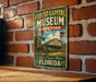 A vintage-style metal sign for the Forest Capital Museum in Florida, featuring an illustration of a building and a wagon wheel.