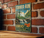 A vintage-style metal sign for Forked Run State Park in Ohio, featuring a scenic landscape with a waterfall and trees.