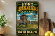 A vintage-style sign for Fort Abraham Lincoln State Park in North Dakota, featuring a cannon and a wooden fence, with a backdrop of a blue sky and clouds.