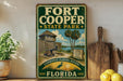 A vintage-style sign for Fort Cooper State Park in Florida, featuring a wooden cabin and palm trees, is displayed on a shelf next to a wooden cutting board and a potted plant.