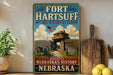 a vintage-style sign for Fort Hartsuff State Historical Park, with a wooden fence and a cannon in the background, along with a potted plant and a wooden cutting board.