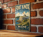 A vintage-style metal sign for the Fort Knox State Historic Site in Maine, featuring an illustration of a fort on a hill with a castle and a sailboat in the background.