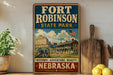 A vintage-style sign for Fort Robinson State Park in Nebraska, featuring a large illustration of a historic building and a cannon, with the text "History, Adventure, Beauty, Nebraska" below.