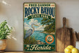 A vintage-style sign for Rocky Bayou State Park in Florida, featuring a heron in the foreground and a scenic landscape with a body of water and trees in the background.