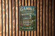 A vintage-style metal sign for Gamble Plantation Historic State Park in Florida, featuring a large illustration of a historic plantation house surrounded by palm trees.