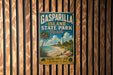 A vintage metal sign for Gasparilla Island State Park in Florida, featuring a beach scene with palm trees and a lighthouse, mounted on a wooden wall.