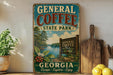 a vintage-style metal sign for General Coffee State Park in Georgia, with a scenic landscape of a lake and trees. The sign is displayed on a shelf, surrounded by various kitchen utensils and a potted plant.