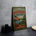 A vintage-style metal sign for Georgia Veterans State Park, featuring an American flag and a scenic landscape, is displayed on a gray countertop next to a stack of black camera lenses.
