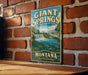 A vintage-style metal sign for Giant Springs State Park in Montana, featuring a scenic image of a waterfall and mountains.