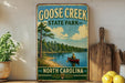 A vintage-style sign for Goose Creek State Park in North Carolina, featuring a scenic image of a person in a canoe on a lake surrounded by pine trees.