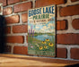 A vintage-style sign for Goose Lake Prairie, Illinois, featuring a scenic landscape with a lake, flowers, and a blue sky with clouds.
