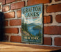 A vintage metal sign for Grafton Lakes State Park, featuring a scenic landscape with a lake and mountains, is displayed on a brick wall.