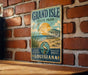 A vintage-style metal sign for Grand Isle State Park in Louisiana, featuring a beach scene with a house and palm trees.