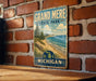 A vintage metal sign for Grand Mere State Park in Michigan, featuring a scenic landscape with a beach, mountains, and a lake.
