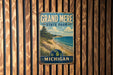 A vintage metal sign for Grand Mere State Park in Michigan, featuring a scenic landscape with a beach, mountains, and a pine tree.