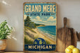 A vintage-style sign for Grand Mere State Park in Michigan, featuring a scenic beach landscape with a pine tree and a wooden fence.
