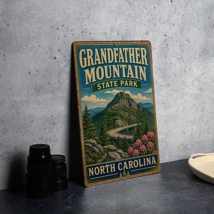 A vintage-style metal sign for Grandfather Mountain State Park in North Carolina, featuring a scenic mountain landscape with a winding road and pink flowers.