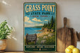 A vintage-style sign for Grass Point State Park in New York, featuring a scenic landscape with a beach, mountains, and pine trees.